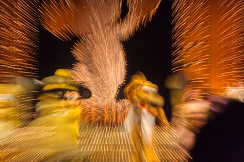 Blurred motion capture of colorful characters amidst a backdrop of vibrant lights, with a central figure outlined by fireworks patterns.