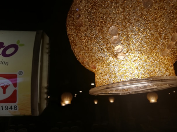 A textured, golden lampshade with circular patterns is prominently featured, illuminated from within. In the background, there's a partially visible advertisement with text and logo elements.