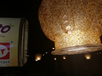 A textured, golden lampshade with circular patterns is prominently featured, illuminated from within. In the background, there's a partially visible advertisement with text and logo elements.