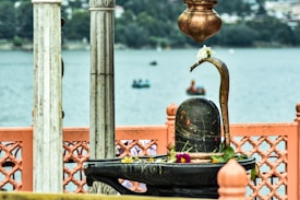 A stone Shiva Lingam adorned with flowers and leaves is situated in a decorative outdoor setting beside a serene water body. Marbled pillars and ornate orange fencing are partially visible. The background features blurred boats on the water and a distant shoreline with lush greenery.