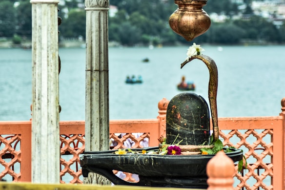 A stone Shiva Lingam adorned with flowers and leaves is situated in a decorative outdoor setting beside a serene water body. Marbled pillars and ornate orange fencing are partially visible. The background features blurred boats on the water and a distant shoreline with lush greenery.