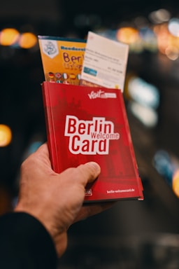 A hand is holding a red Berlin Welcome Card along with some tourist brochures against a blurred background with bright lights. The card features the Berlin skyline in silhouette.