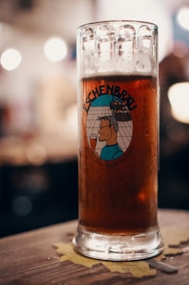 A glass of dark beer with foam at the top sits on a wooden table with a coaster underneath. The glass features a cartoon illustration with the word 'Eschenbräu' prominently displayed. The background is softly blurred, suggesting a cozy, dimly lit atmosphere.