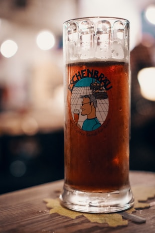 A glass of dark beer with foam at the top sits on a wooden table with a coaster underneath. The glass features a cartoon illustration with the word 'Eschenbräu' prominently displayed. The background is softly blurred, suggesting a cozy, dimly lit atmosphere.