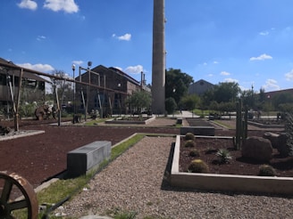A landscaped garden area with neatly arranged pathways and garden beds. The centerpiece is a tall, cylindrical chimney. Industrial style buildings can be seen in the background. The pathways are bordered by greenery and various small desert plants.