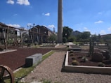 This image depicts an industrial-style garden or park with a tall chimney-like structure in the center. The landscape includes manicured garden beds with stones, gravel paths, and various desert plants such as cacti. In the background, there are industrial buildings with visible metal pipes and machinery, giving it a historic factory or mill appearance. The sky is clear and blue with a few scattered clouds.