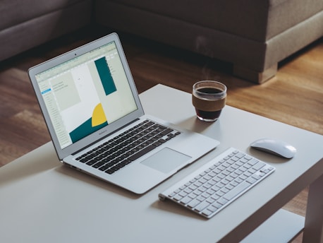 A modern workspace featuring an open laptop displaying a design application on a white desk. Accompanying the laptop are a wireless keyboard and mouse, along with a steaming cup of coffee. The setting appears cozy with a comfortable brown sofa in the background and a wooden floor.