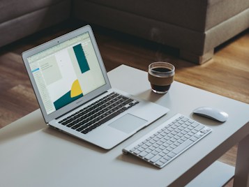 A modern workspace featuring an open laptop displaying a design application on a white desk. Accompanying the laptop are a wireless keyboard and mouse, along with a steaming cup of coffee. The setting appears cozy with a comfortable brown sofa in the background and a wooden floor.