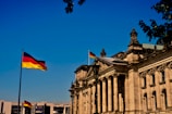 A grand historic building with ornate architectural details and tall columns, displaying the German flag on top. The structure is bathed in sunlight against a clear blue sky. A second German flag is seen prominently on a flagpole in the foreground. Leafy tree branches partially frame the upper part of the image.