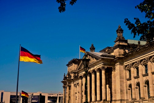 A grand historic building with ornate architectural details and tall columns, displaying the German flag on top. The structure is bathed in sunlight against a clear blue sky. A second German flag is seen prominently on a flagpole in the foreground. Leafy tree branches partially frame the upper part of the image.