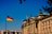 A grand historic building with ornate architectural details and tall columns, displaying the German flag on top. The structure is bathed in sunlight against a clear blue sky. A second German flag is seen prominently on a flagpole in the foreground. Leafy tree branches partially frame the upper part of the image.