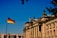 A grand historic building with ornate architectural details and tall columns, displaying the German flag on top. The structure is bathed in sunlight against a clear blue sky. A second German flag is seen prominently on a flagpole in the foreground. Leafy tree branches partially frame the upper part of the image.