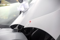 A close-up of a white car with the hood prominently displayed. The car features a badge that reads '720S' in red lettering. A white ribbon is attached to the side mirror, suggesting it may be dressed for a special occasion.