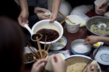 A group of people is preparing dumplings on a table filled with various ingredients in bowls. A hand is pouring a dark sauce from a large bowl using a ladle. Several hands are holding onto chopsticks, picking up wrappers and adding fillings. Bowls contain ingredients like minced meat and vegetables, and there are small dishes with condiments.