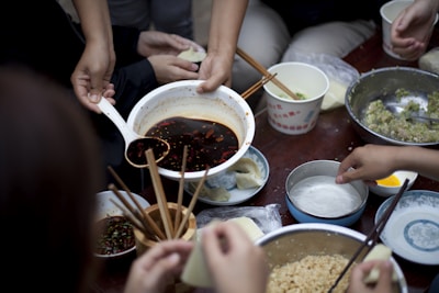 A group of people is preparing dumplings on a table filled with various ingredients in bowls. A hand is pouring a dark sauce from a large bowl using a ladle. Several hands are holding onto chopsticks, picking up wrappers and adding fillings. Bowls contain ingredients like minced meat and vegetables, and there are small dishes with condiments.