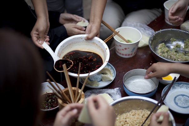 A group of people is preparing dumplings on a table filled with various ingredients in bowls. A hand is pouring a dark sauce from a large bowl using a ladle. Several hands are holding onto chopsticks, picking up wrappers and adding fillings. Bowls contain ingredients like minced meat and vegetables, and there are small dishes with condiments.
