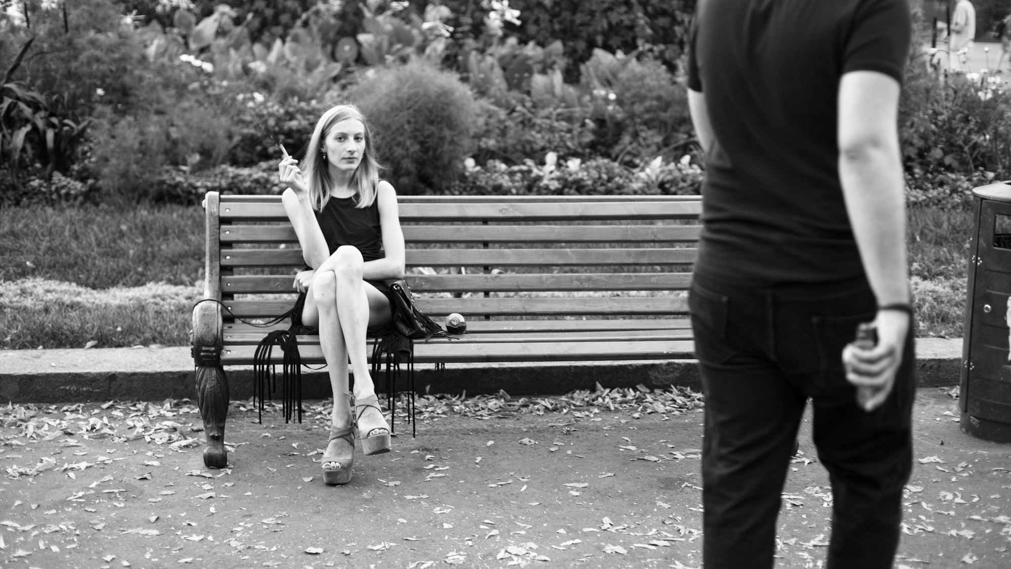 A chic cool woman in an LBD sitting on a bench in a park, smoking.