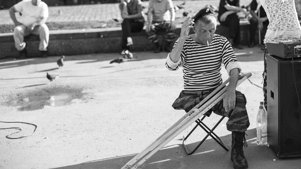 Photo en niveaux de gris d’un homme assis sur un tabouret pliant avec des béquilles devant des gens