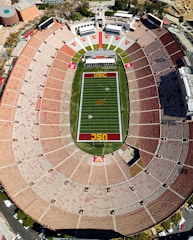 An aerial view of a large, oval-shaped stadium featuring a green field marked for American football, with the letters 'USC' prominently displayed. The seating is mostly empty and consists of orange benches arranged in sections. Surrounding structures and roadways are visible around the stadium.