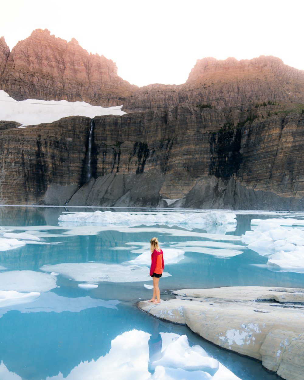 woman in red and orange sweater stands on stone beside water