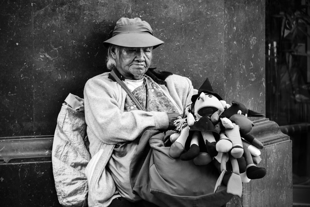 grayscale photography of man carrying plush toys while sitting against wall