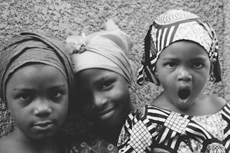 Three children are standing in front of a textured wall. They are wearing headscarves and patterned clothing. One child is making a playful expression with an open mouth, while the others have subtle smiles.
