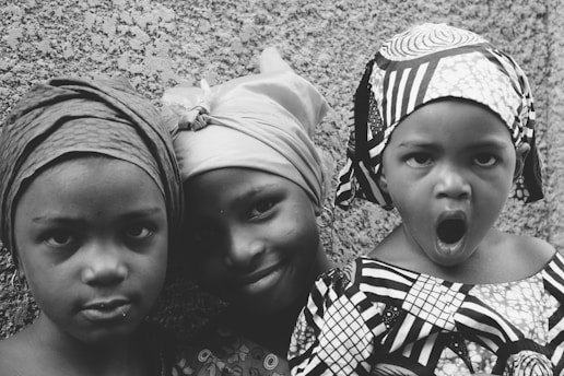 Three children are standing in front of a textured wall. They are wearing headscarves and patterned clothing. One child is making a playful expression with an open mouth, while the others have subtle smiles.