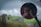 A photograph taken through the side mirror of a vehicle, capturing a person holding a camera up to their face. The background reveals a blurred outdoor scene with green fields, a road, some fencing, power lines, and a person riding a motorcycle in the distance under a cloudy sky.
