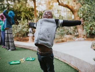 A joyful child with open arms is playing outside on green grass, expressing excitement and happiness. Other children are visible in the background, engaged in playful activities. Trees and a wooden path add a natural setting.