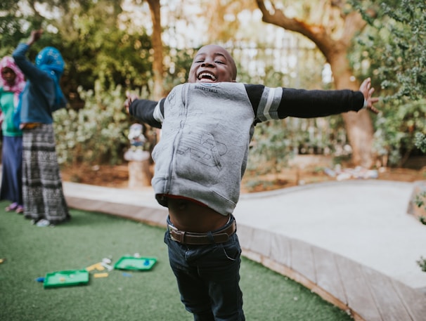A joyful child with open arms is playing outside on green grass, expressing excitement and happiness. Other children are visible in the background, engaged in playful activities. Trees and a wooden path add a natural setting.