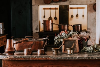 A rustic kitchen setting featuring copper pots and utensils placed on a wooden counter. Bunches of grapes and dried flowers are arranged decoratively around an old slate. The background displays hanging kitchen tools against stone walls, creating a warm, vintage atmosphere.