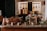 A rustic kitchen setting featuring copper pots and utensils placed on a wooden counter. Bunches of grapes and dried flowers are arranged decoratively around an old slate. The background displays hanging kitchen tools against stone walls, creating a warm, vintage atmosphere.