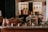 A rustic kitchen setting featuring copper pots and utensils placed on a wooden counter. Bunches of grapes and dried flowers are arranged decoratively around an old slate. The background displays hanging kitchen tools against stone walls, creating a warm, vintage atmosphere.