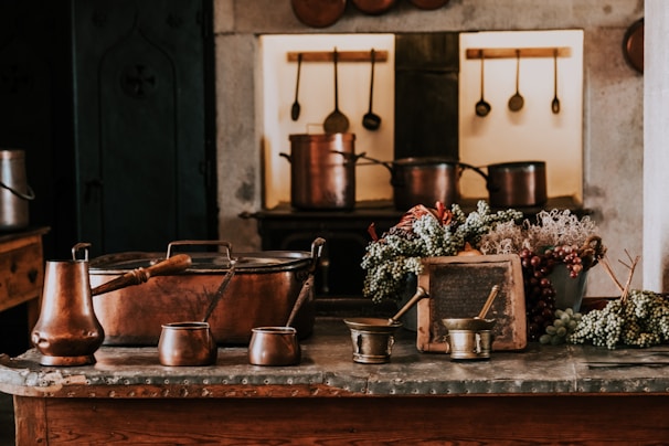 A rustic kitchen setting featuring copper pots and utensils placed on a wooden counter. Bunches of grapes and dried flowers are arranged decoratively around an old slate. The background displays hanging kitchen tools against stone walls, creating a warm, vintage atmosphere.
