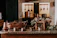 A rustic kitchen setting featuring copper pots and utensils placed on a wooden counter. Bunches of grapes and dried flowers are arranged decoratively around an old slate. The background displays hanging kitchen tools against stone walls, creating a warm, vintage atmosphere.