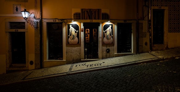 An exterior view of a building at night with illuminated artwork of dancers on either side of the entrance. The building has a sign above the door with the word 'Foxtrot.' The narrow street in front is paved with cobblestones and slopes down to the right.