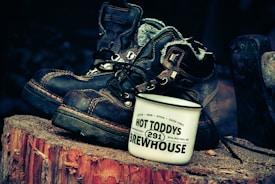 A pair of rugged, well-worn boots is placed on a wooden stump, accompanied by an enamel mug. The mug is white and prominently features the text 'Hot Toddys Brewhouse.' The overall ambiance suggests a rustic, outdoorsy setting.