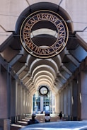 An architectural view featuring an arched walkway with a prominent sign for the Federal Reserve Bank of San Francisco above. The passageway is flanked by tall columns and leads towards a windowed building in the background. There is a person wearing dark clothing and a mask visible at the bottom, along with a car partially visible. The lighting creates patterns of shadow and light along the walkway.