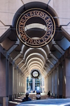 An architectural view featuring an arched walkway with a prominent sign for the Federal Reserve Bank of San Francisco above. The passageway is flanked by tall columns and leads towards a windowed building in the background. There is a person wearing dark clothing and a mask visible at the bottom, along with a car partially visible. The lighting creates patterns of shadow and light along the walkway.