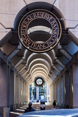 An architectural view featuring an arched walkway with a prominent sign for the Federal Reserve Bank of San Francisco above. The passageway is flanked by tall columns and leads towards a windowed building in the background. There is a person wearing dark clothing and a mask visible at the bottom, along with a car partially visible. The lighting creates patterns of shadow and light along the walkway.