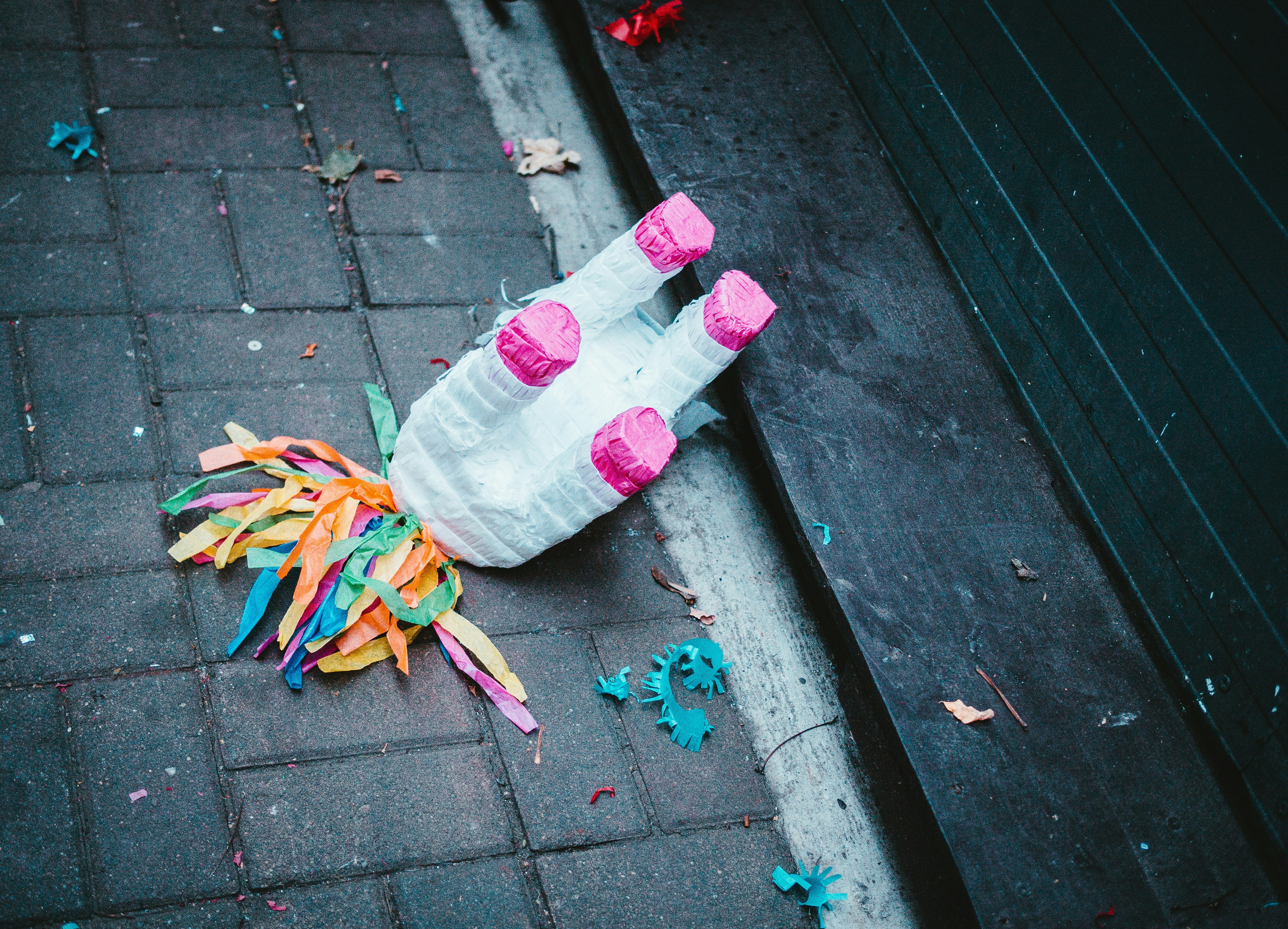 A white animal piñata with a rainbow streamer tail and pink feet is upside down in front of a dark gray door.