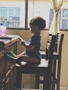 A young child sits on a wooden chair by a table, focused on a smartphone held in both hands. The child is wearing a blue shirt and green shoes. The room appears to be softly lit by natural light coming through a window with sheer curtains, adding warmth to the setting. A pink cup rests on the table.