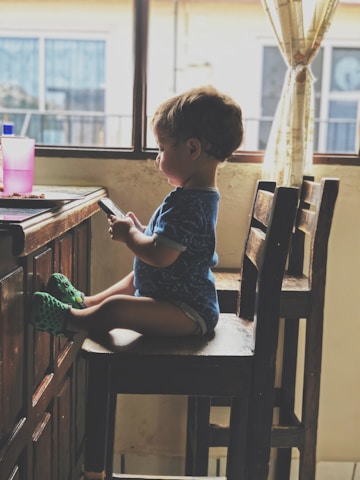 A young child sits on a wooden chair by a table, focused on a smartphone held in both hands. The child is wearing a blue shirt and green shoes. The room appears to be softly lit by natural light coming through a window with sheer curtains, adding warmth to the setting. A pink cup rests on the table.