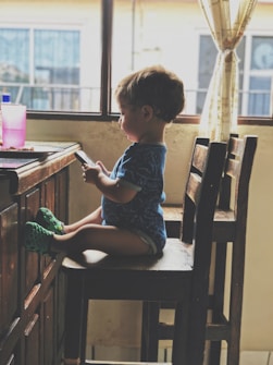 A young child sits on a wooden chair by a table, focused on a smartphone held in both hands. The child is wearing a blue shirt and green shoes. The room appears to be softly lit by natural light coming through a window with sheer curtains, adding warmth to the setting. A pink cup rests on the table.