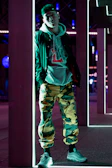 Model wearing a tech-wear jacket standing against a dark urban wall lit by green neon.