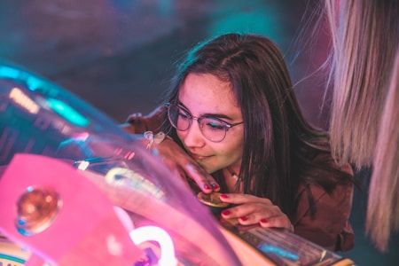 A young woman with glasses is closely examining an arcade or pinball machine, focused on the game. The machine features colorful lights and a reflective surface, creating a vibrant and dynamic atmosphere.