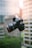 A DSLR camera is captured in mid-air with a blurred urban background, featuring a modern skyscraper with reflective glass windows. The camera's details, such as buttons and lens, are clearly visible.