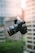 A DSLR camera is captured in mid-air with a blurred urban background, featuring a modern skyscraper with reflective glass windows. The camera's details, such as buttons and lens, are clearly visible.