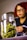 A glass jar filled with green cannabis buds sits prominently in the foreground. In the background, a person with red hair and glasses appears blurred.