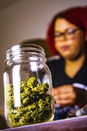 A glass jar filled with green cannabis buds sits prominently in the foreground. In the background, a person with red hair and glasses appears blurred.